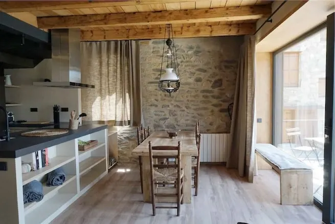 Sala con cocina, mesa de comedor, techo de vigas de madera y pared de piedra
